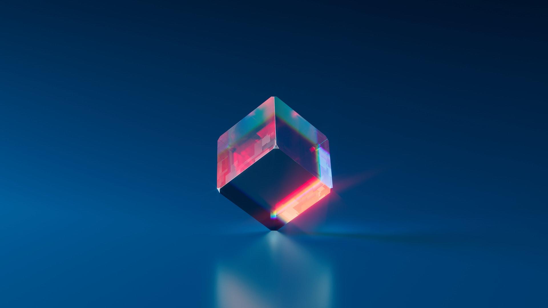A computer 3D graphic rendering of a colorful crystal cube on a blue background.