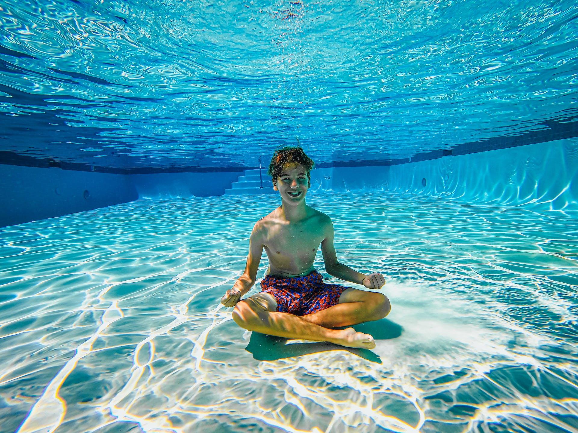 Underwater photo of a child confidently sitting cross-legged at the bottom of a pool and smiling.