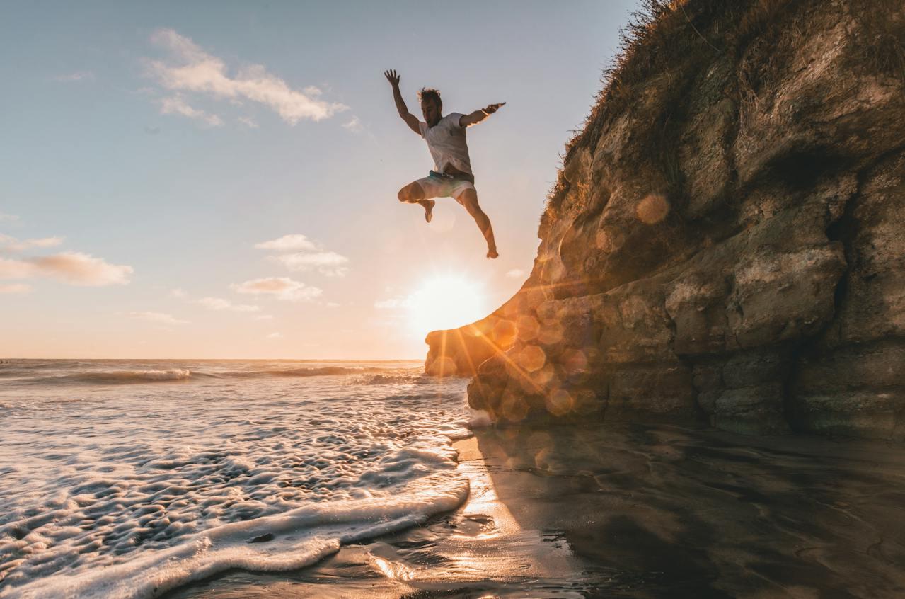 A man jumps from a cliff into the ocean at sunset.