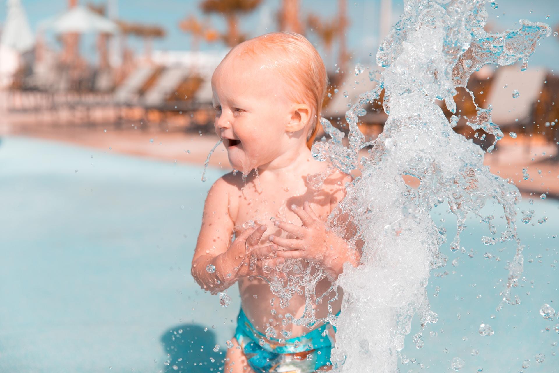 A toddler standing in shallow water getting splashed and laughing.