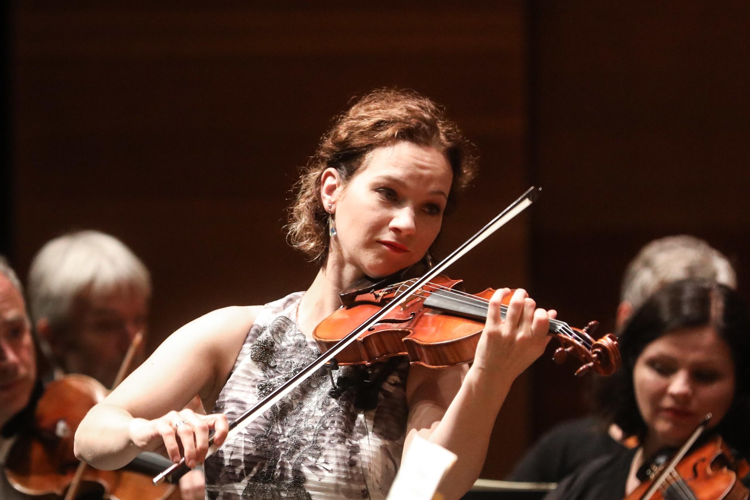 A woman playing the violin on stage with an orchestra in the background.