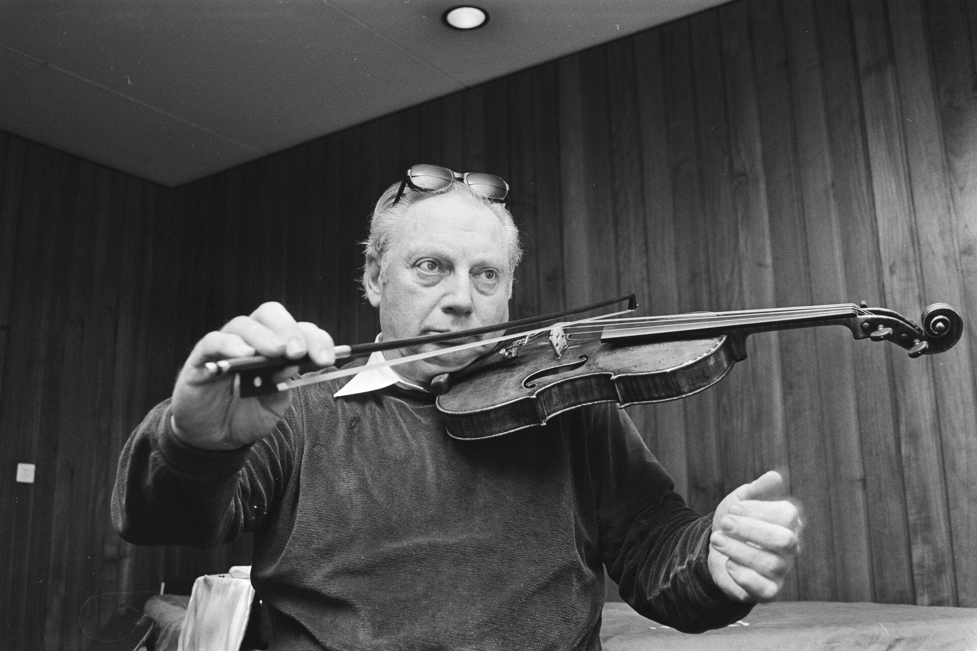 Black and white photo of a man playing the violin indoors, wearing glasses on his head.