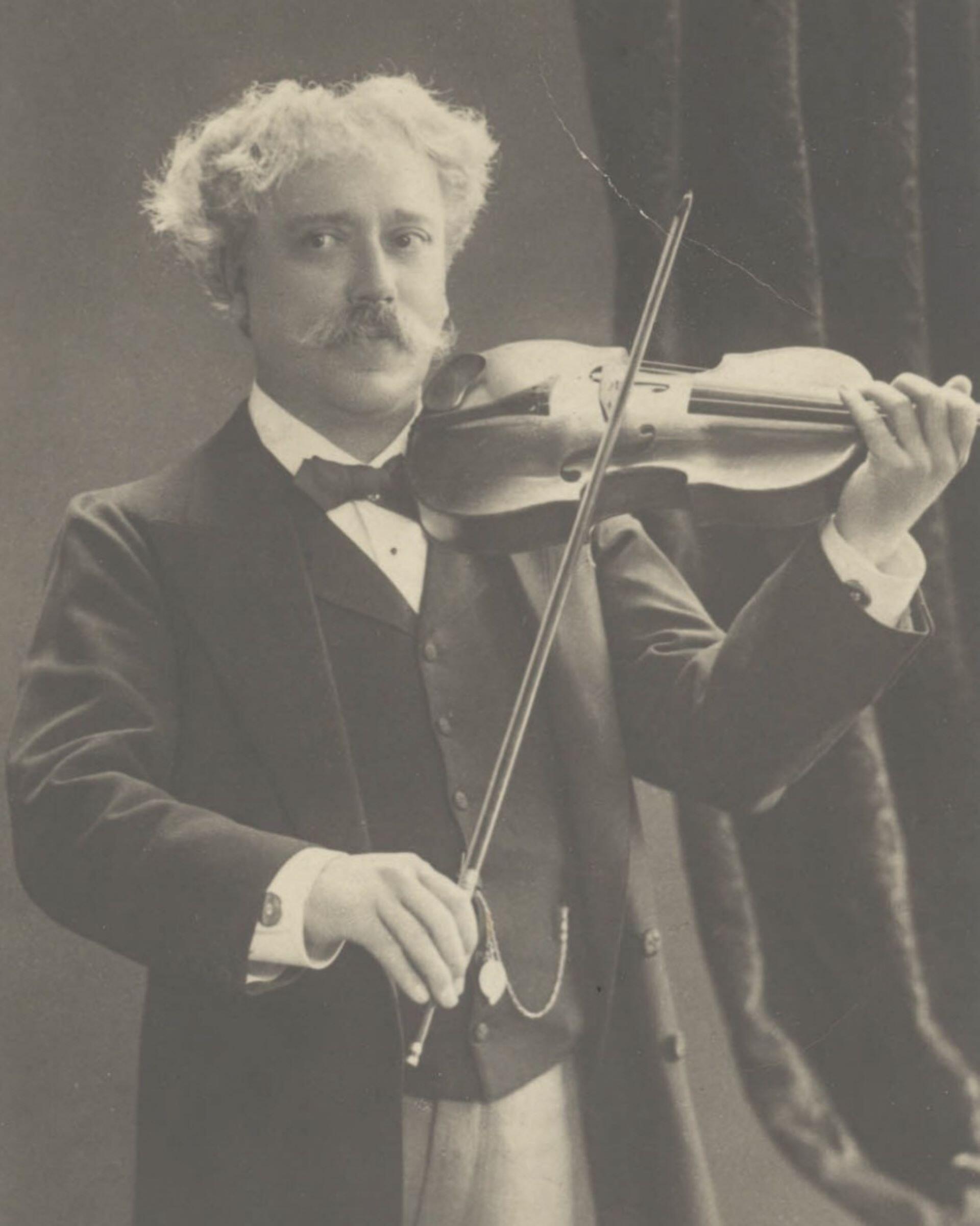 Black and white portrait of a man with a moustache holding a violin and bow in a formal pose.
