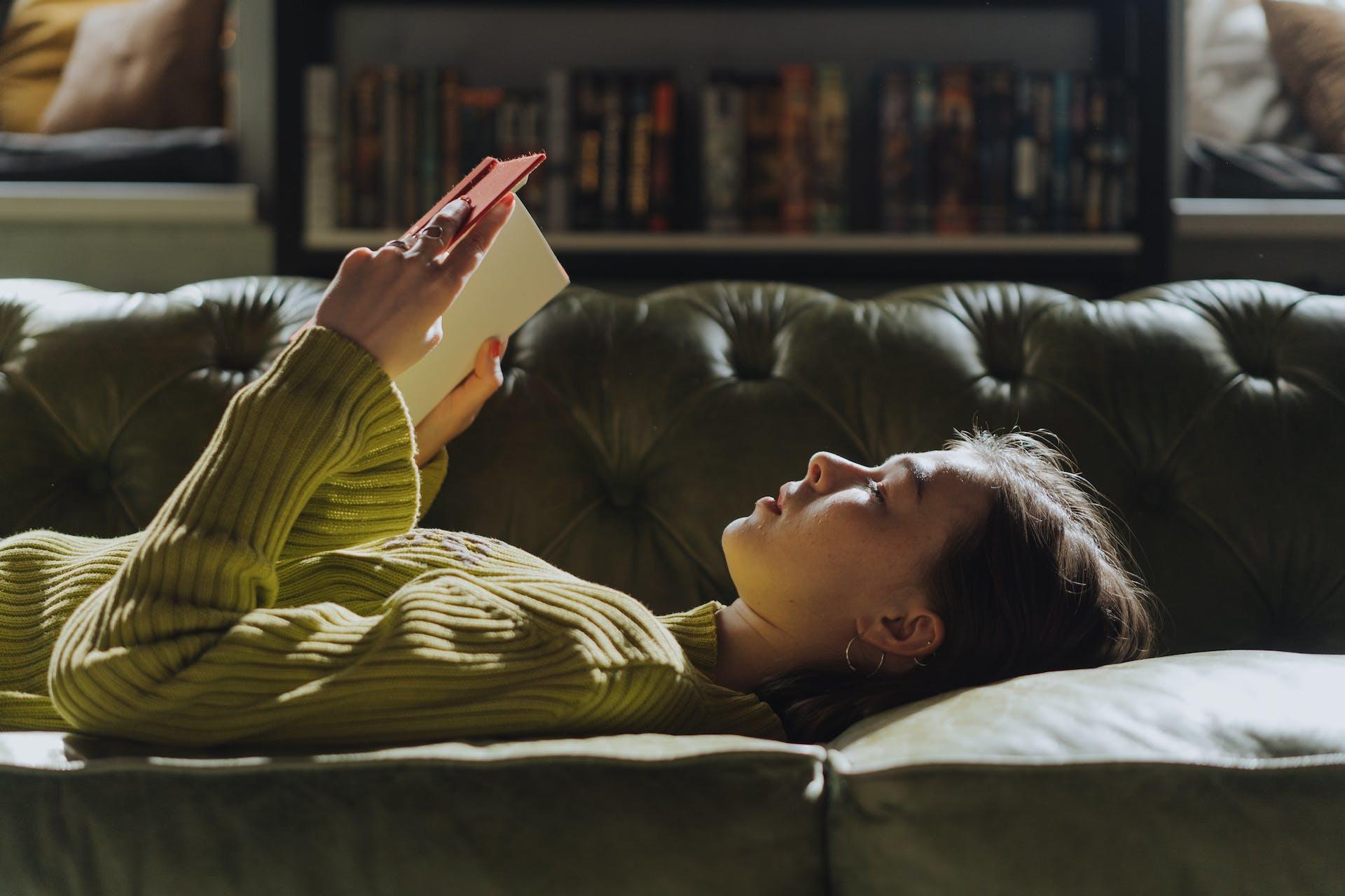 A woman lying down on a couch, reading a book.