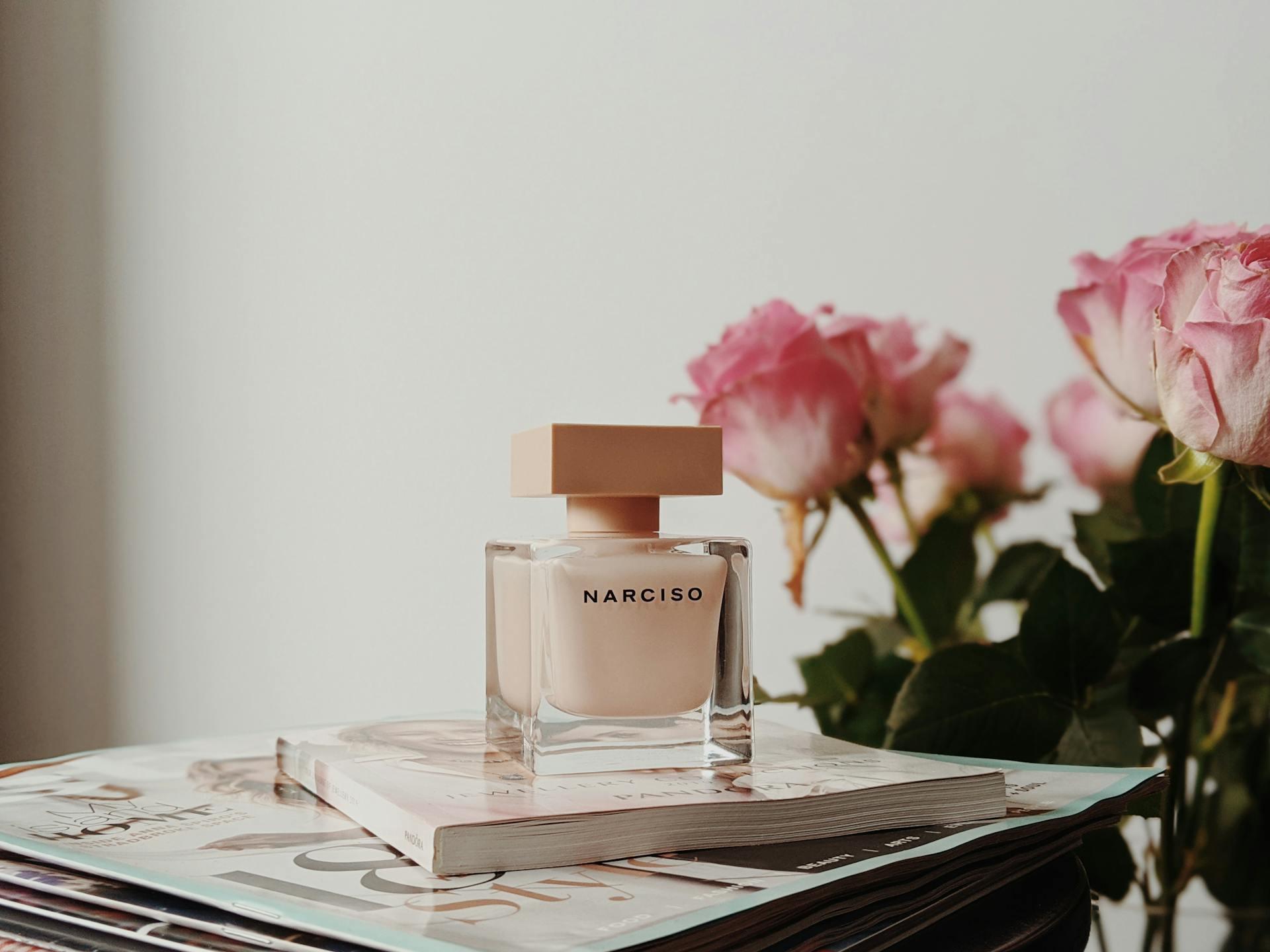 photo of a blush pink perfume in a clear glass bottle with a matte pink top, on top of pastel pink magazines and in front of pink roses