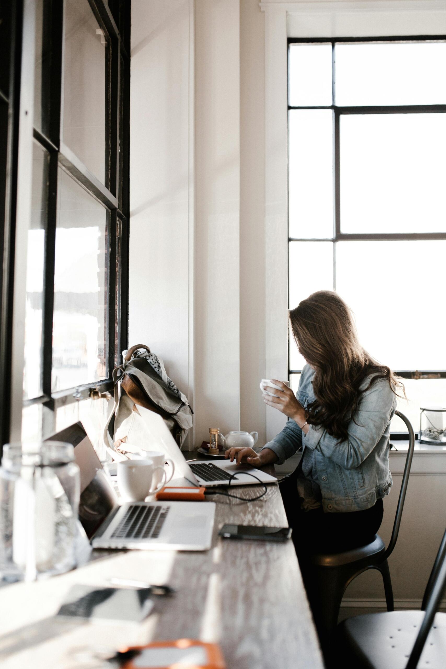 Woman in a gray jacket sitting beside a desk, looking at a laptop screen with focus.