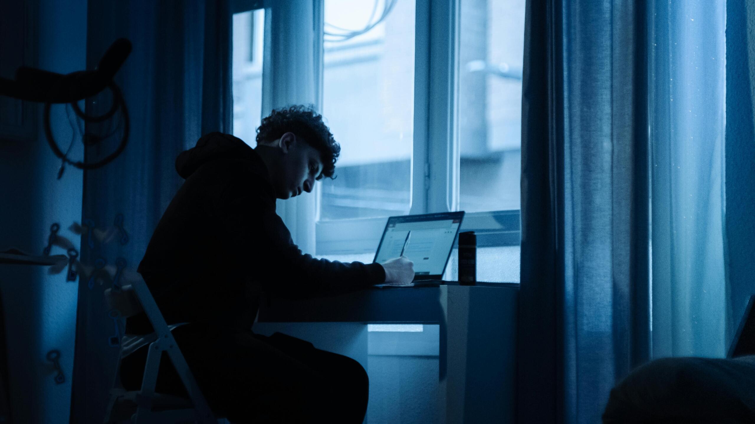 Young man typing on a laptop by a window at night, illuminated by screen light and city lights outside.