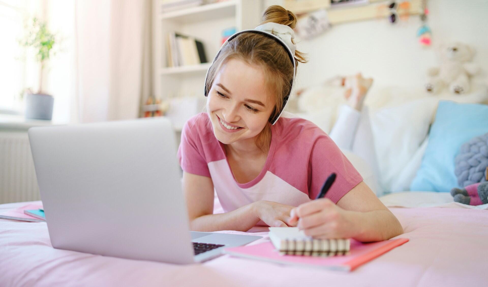 A girl in a pink shirt laying down on her bed in front of a computer.
