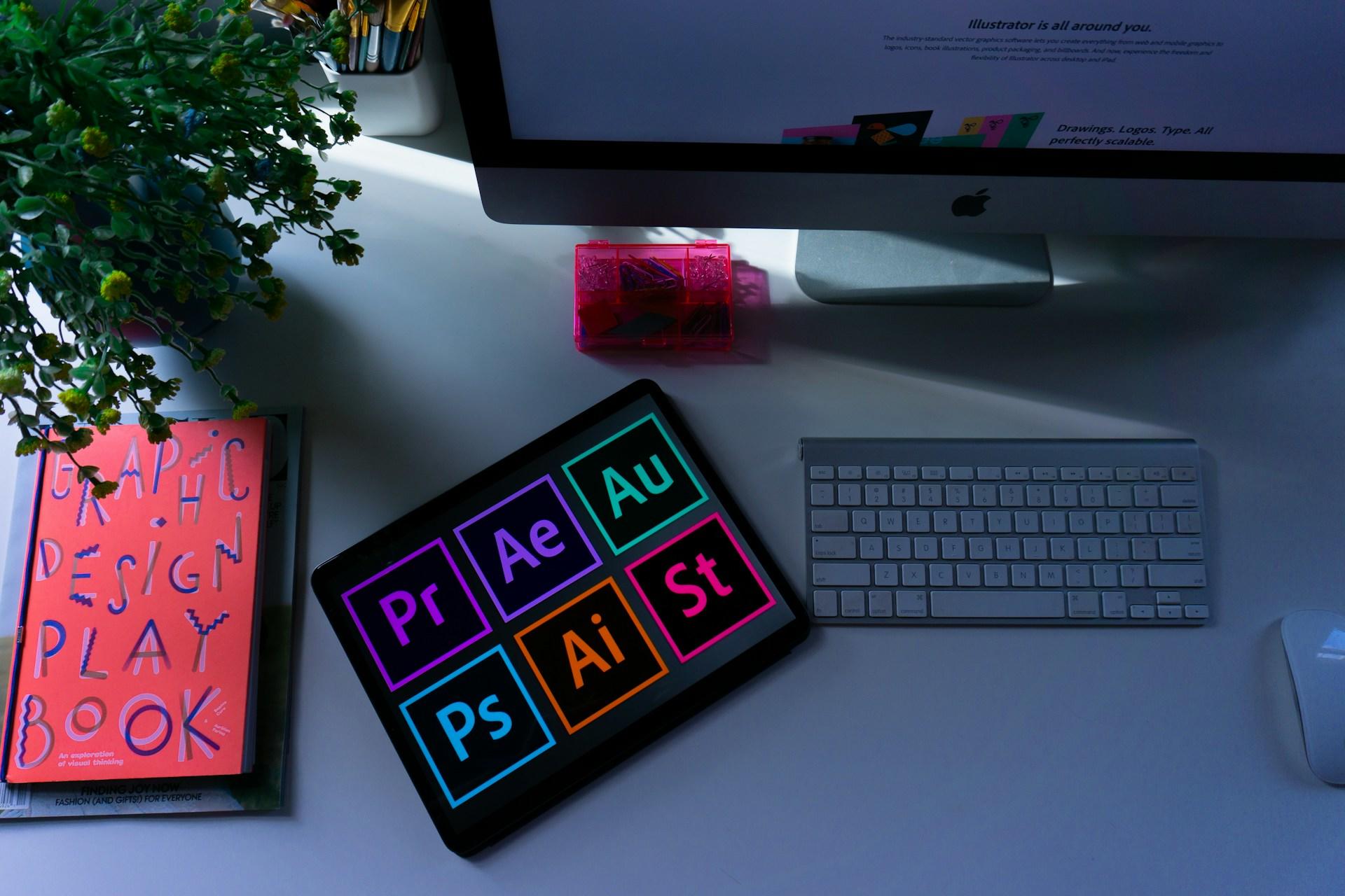 A tablet showing the logos of a variety of Adobe products, near some graphic design books and a desktop computer