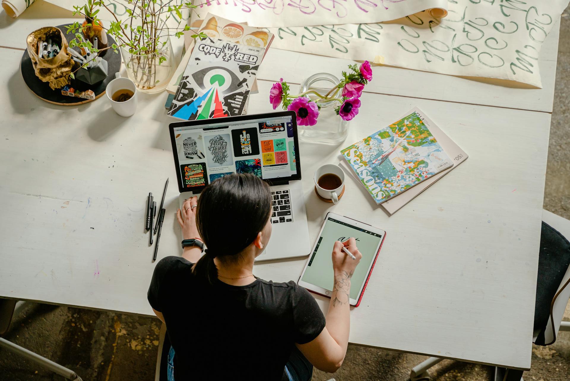 A graphic designer researching designs and making sketches using a laptop, magazines, and an iPad.