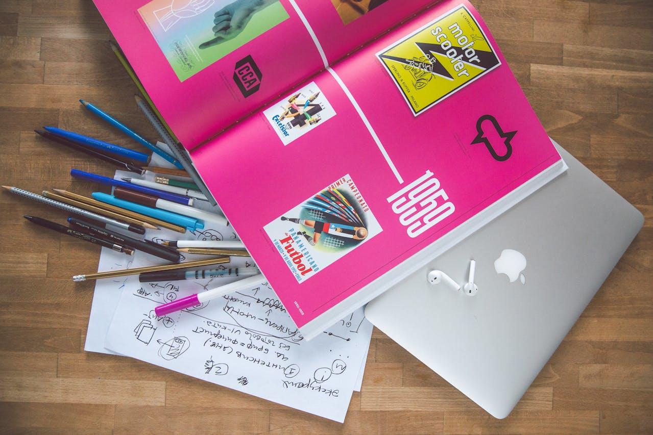 A top-down view of a graphic design history book with a magenta page open on top of a closed laptop with lots of pens and a page of notes nearby.