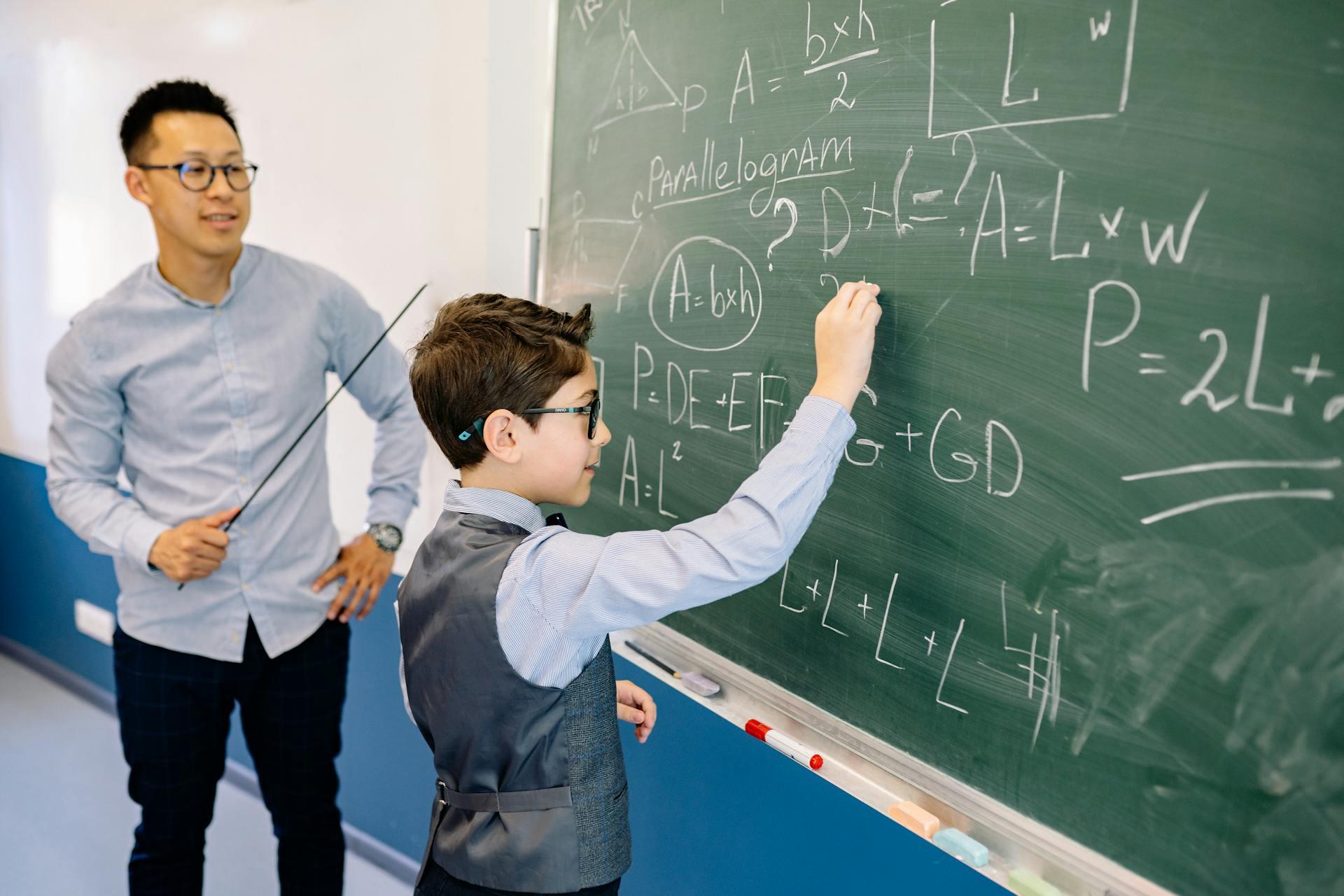 A young student solving a math problem on a chalkboard while his teacher watches