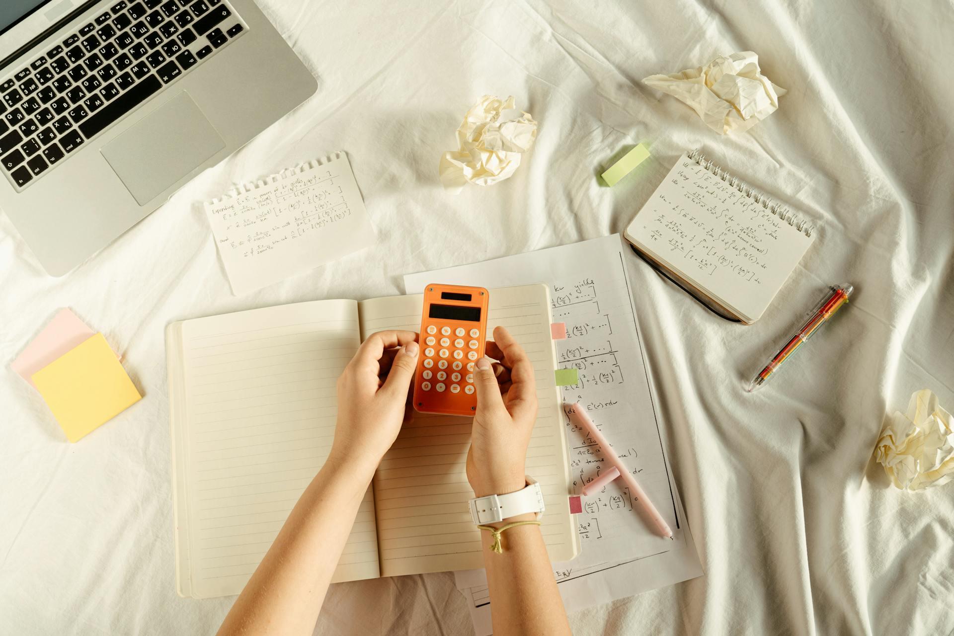 A notebook open on top of math class notes with math problems written on small papers nearby. A hand holds an orange calculator.