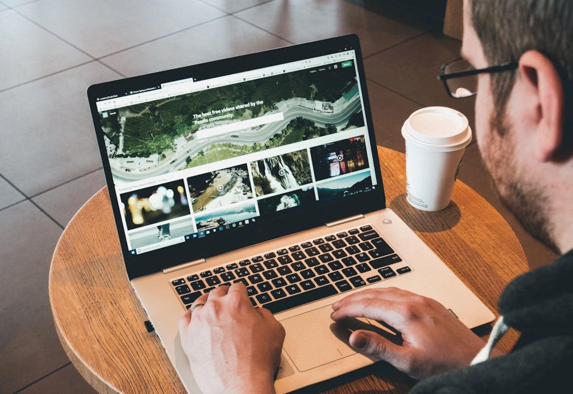 Man at a desk with a coffee cup, looking intently at a laptop screen displaying a design video.