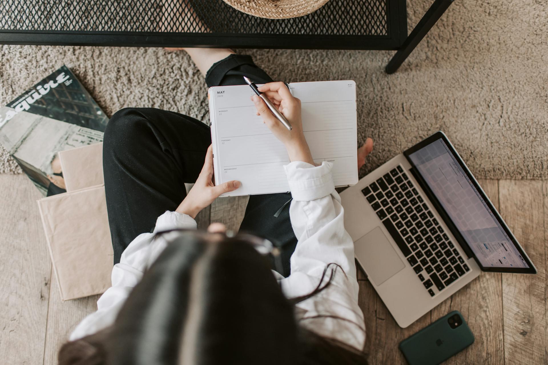 A girl sitting on the flow with a planner in hand and her computer on the ground;
