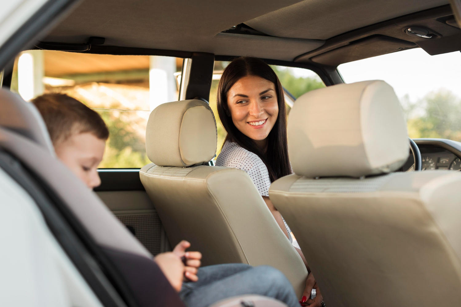 A young woman in the front seat of the car, looking and smiling at a child in a booster seat in the back seat