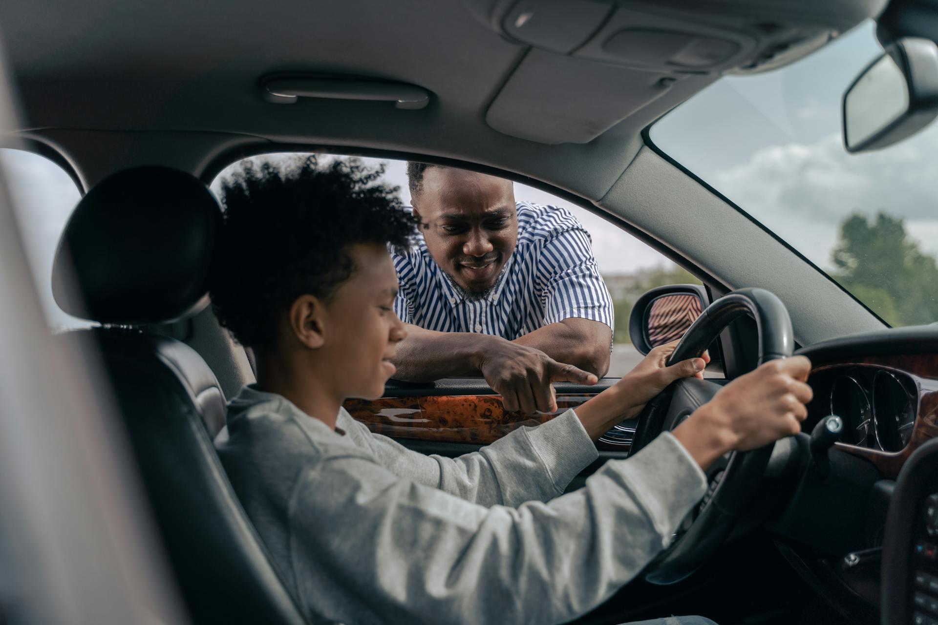 A father helps his teen son learn about driving by pointing out the features near the steering wheel