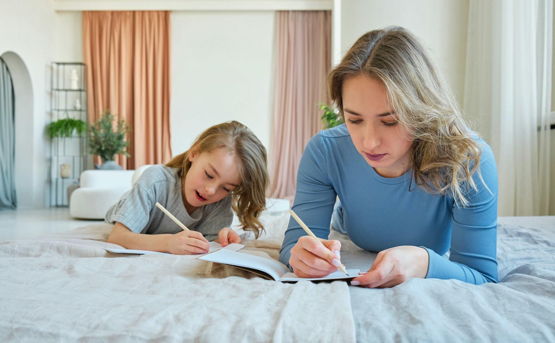 A lady and a young boy laying on a bed writing in a notebook.