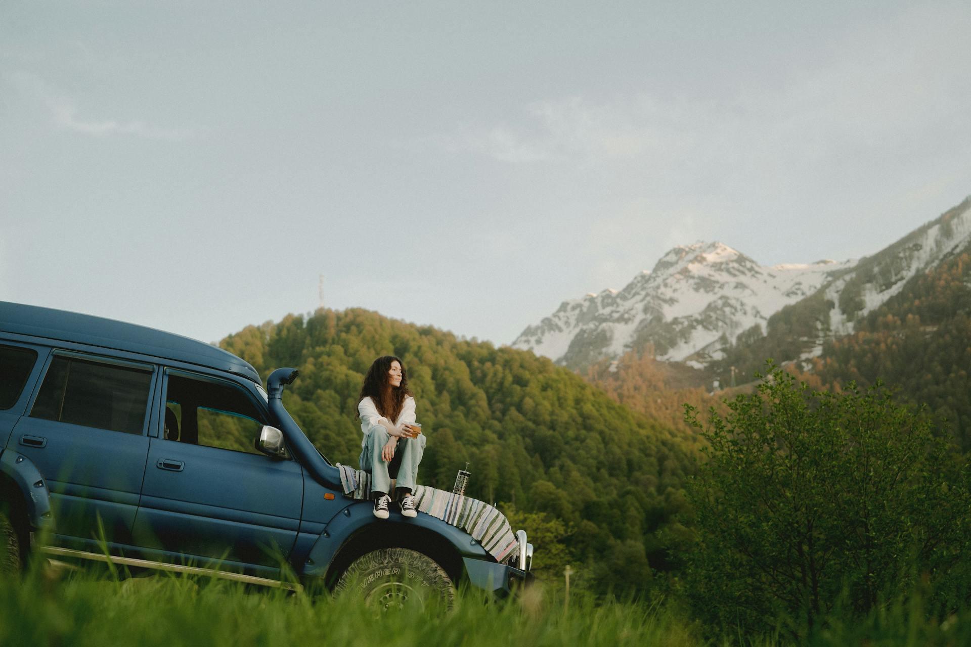 A young woman sitting on the hood of an SUV near some mountains