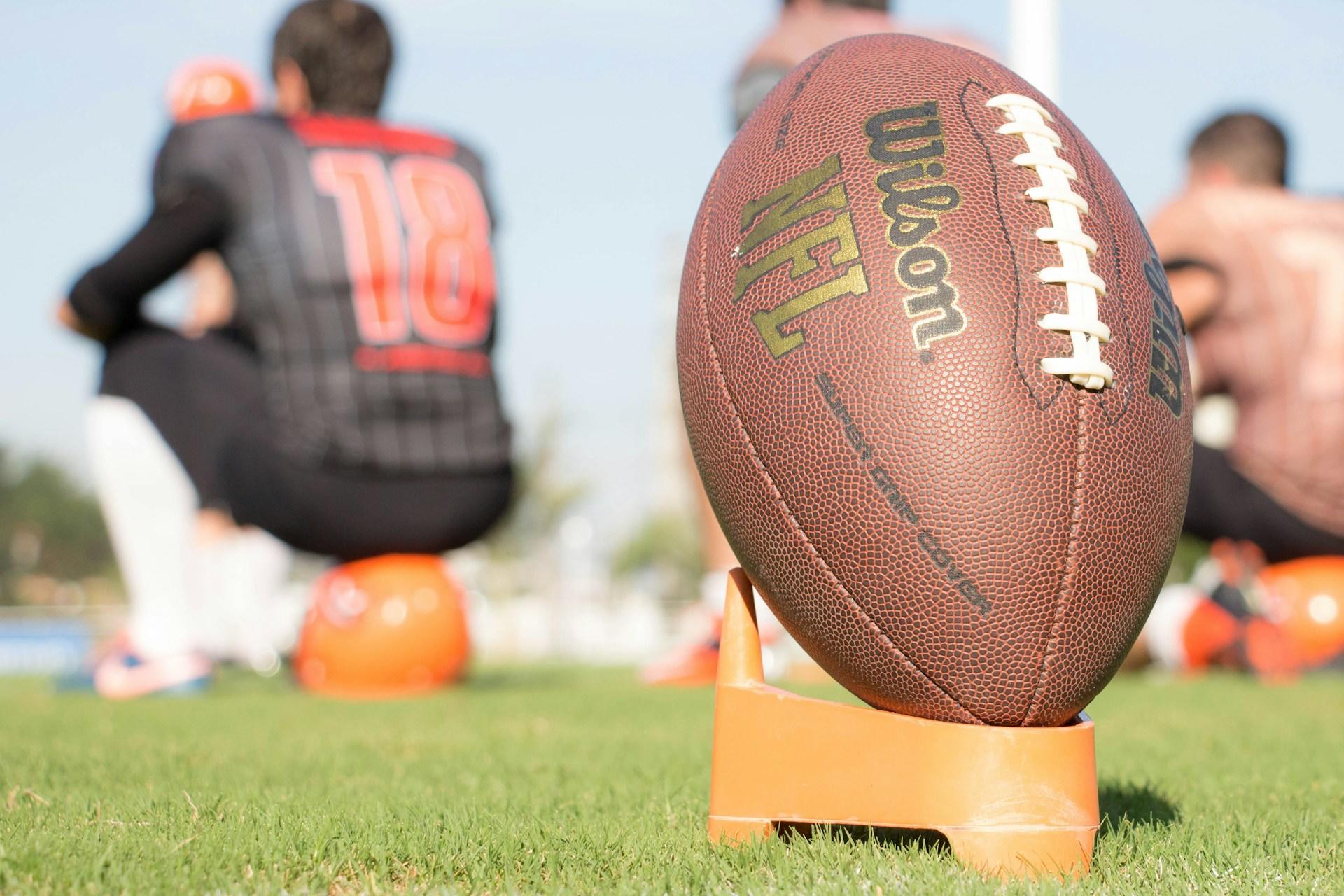 An NFL official football in a tee on a football field with a player in the background