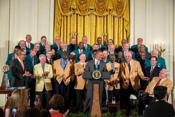 The former Miami Dolphins players from the 1972 team standing behind Obama as he stands at a podium delivering a speech