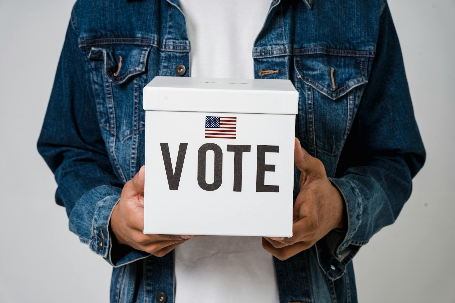 a person holding a small white box with an American flag sticker and large word "vote" on it