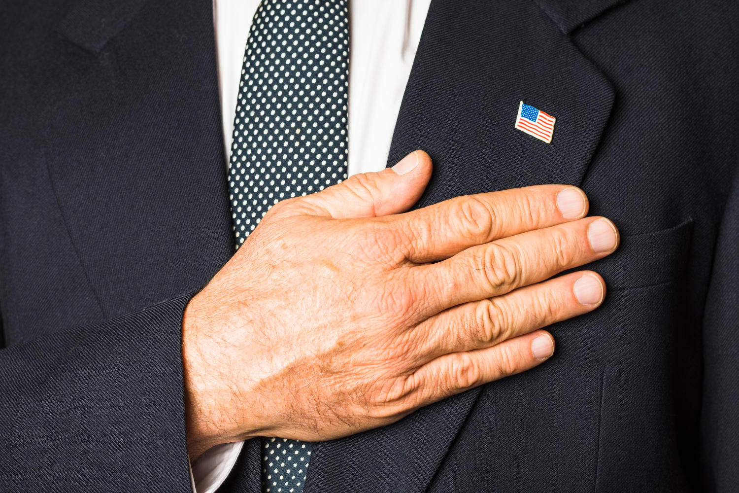 A hand on the breast of a suit with an American flag lapel pin