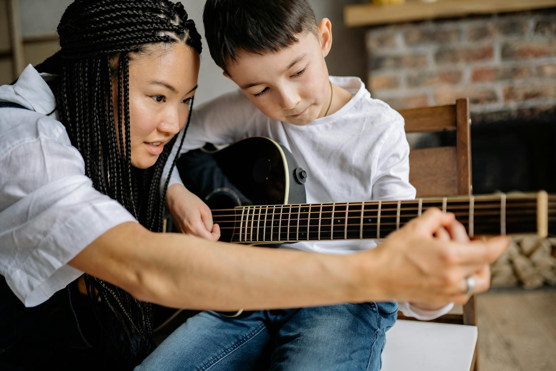 A young boy learning to play guitar while being helped with finger positions by a tutor.