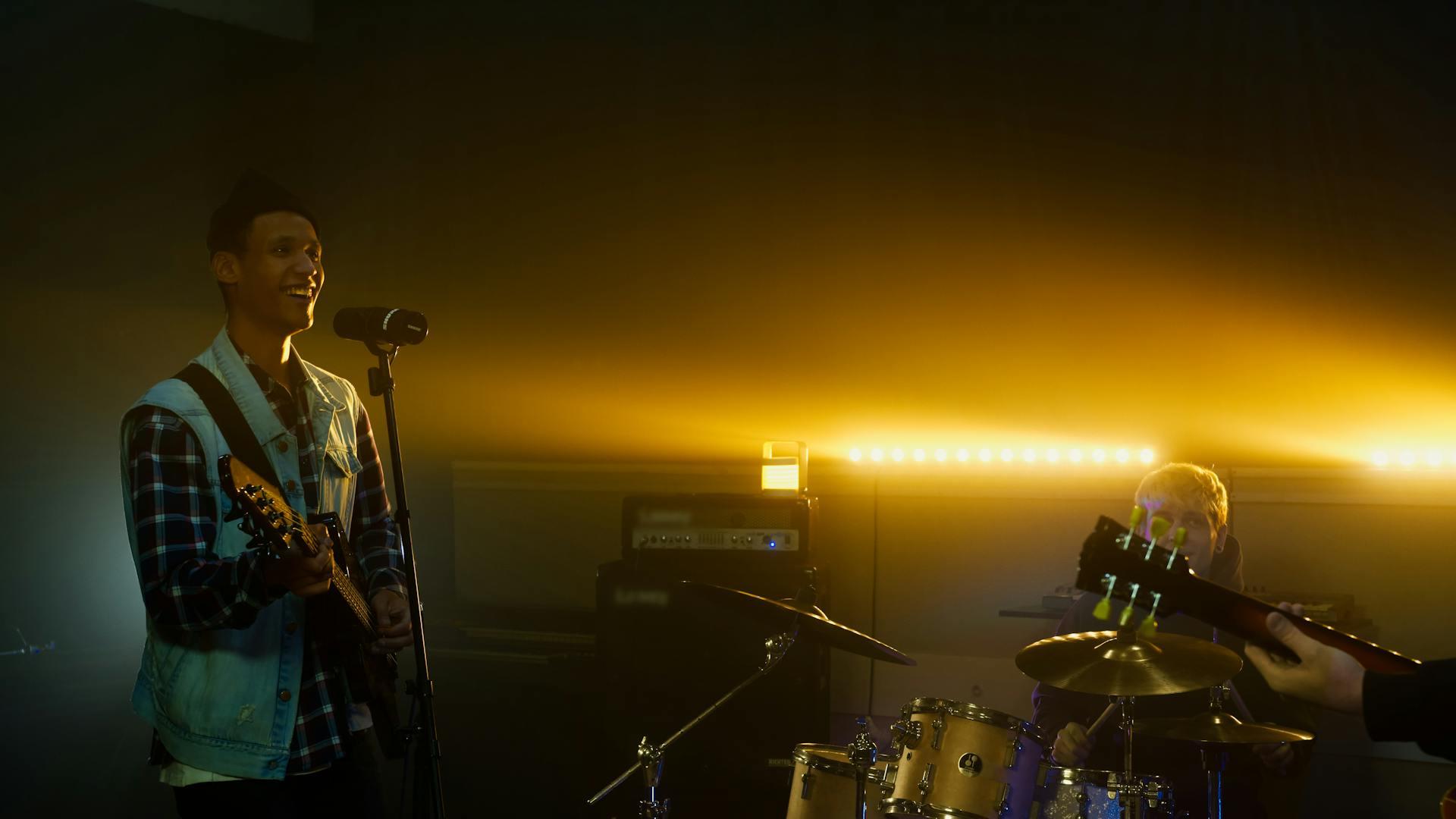 A man singing into a microphone on the left, with bandmates including drummer and guitarist on stage with him.