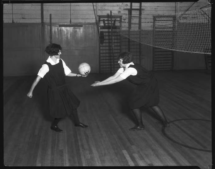 Two women dressed in black dresses with white shirts underneath (1920s PE uniform) playing volleyball in a gymnasium