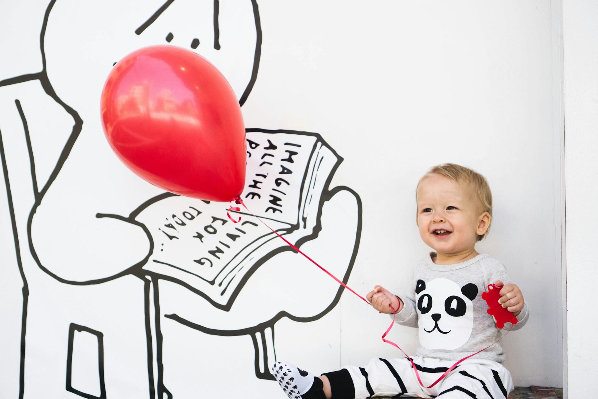 A toddler playing with a red balloon.