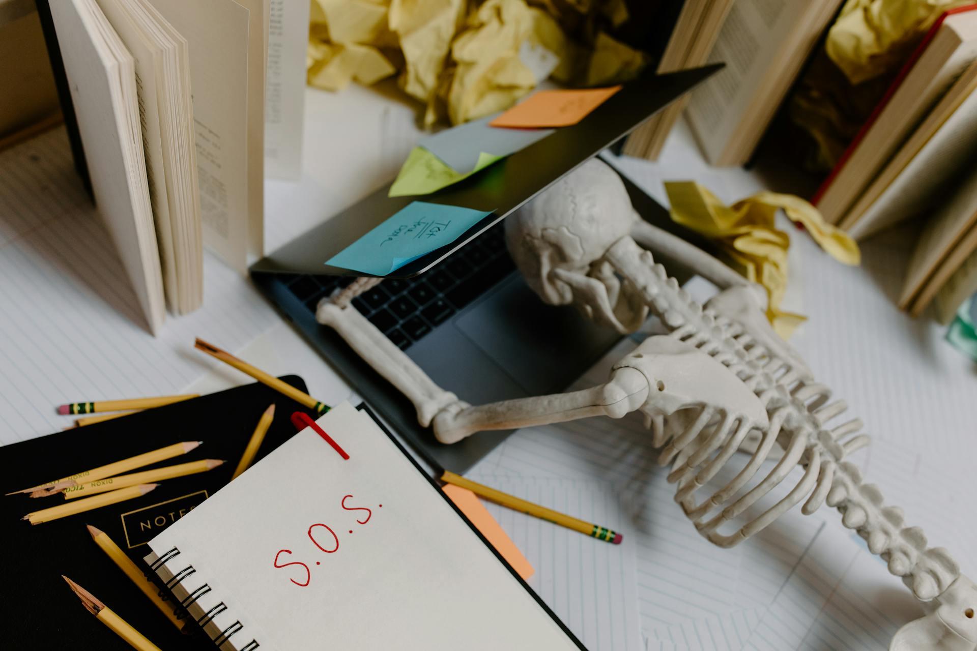 A plastic skeleton face-down on a laptop with a note nearby that reads "S.O.S."