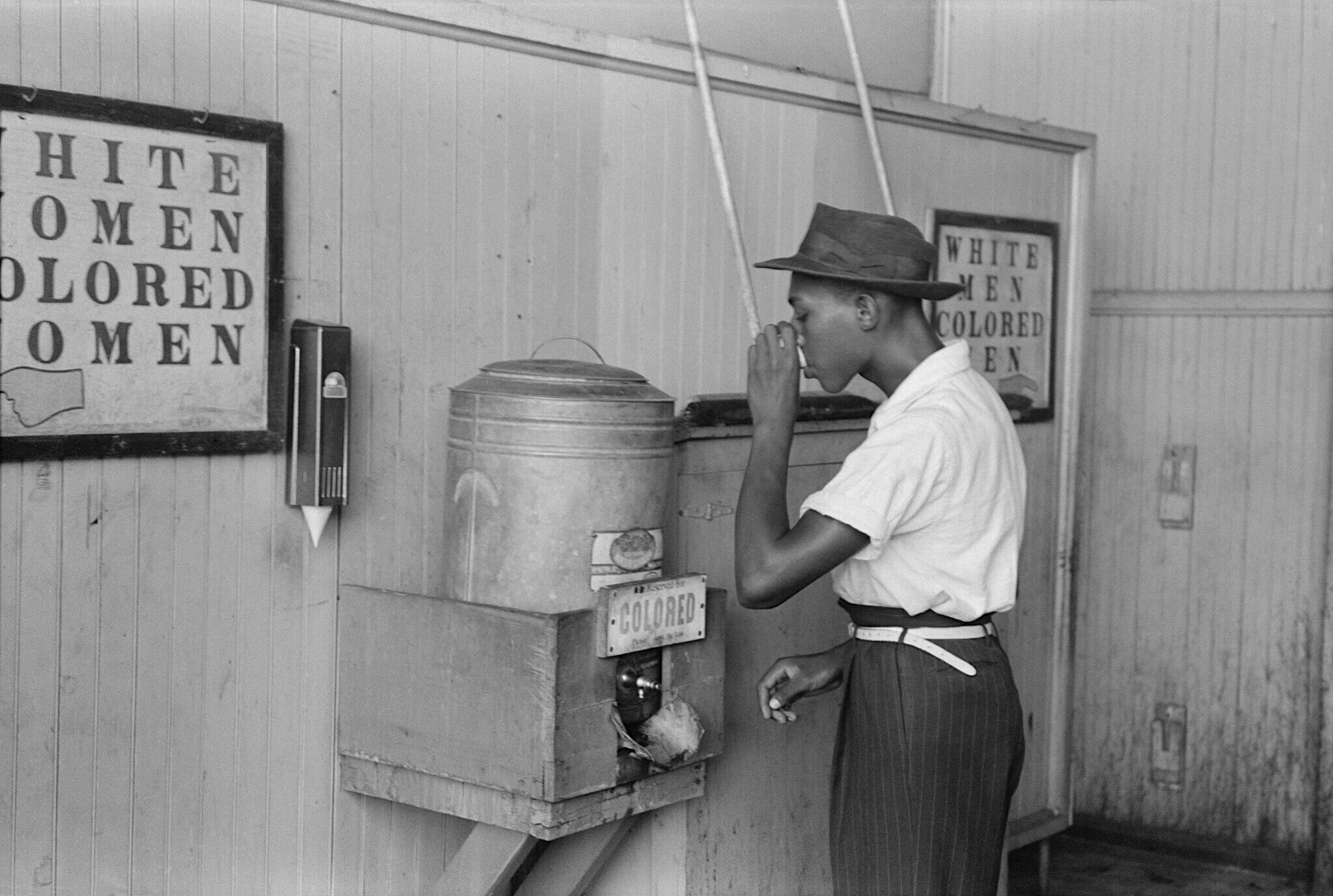 A Black person drinks from a water cooler labelled "Colored" in a black and white photo from 1939.