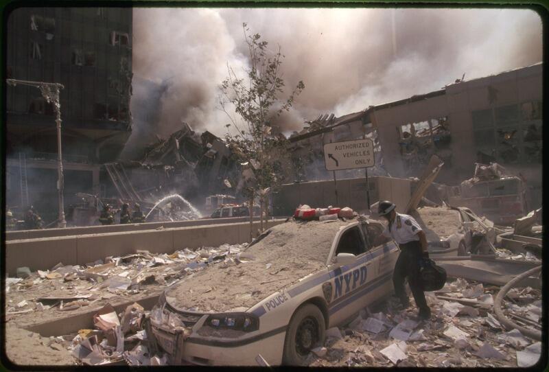 A photo of a police car and the surrounding ground covered in a thick layer of dust and debris. In the background, firefighters are spraying the rubble of a collapsed building.