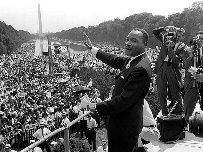 MLK standing on a stage at the March on Washington, a crowd of thousands of peaceful protesters stands on the lawn.