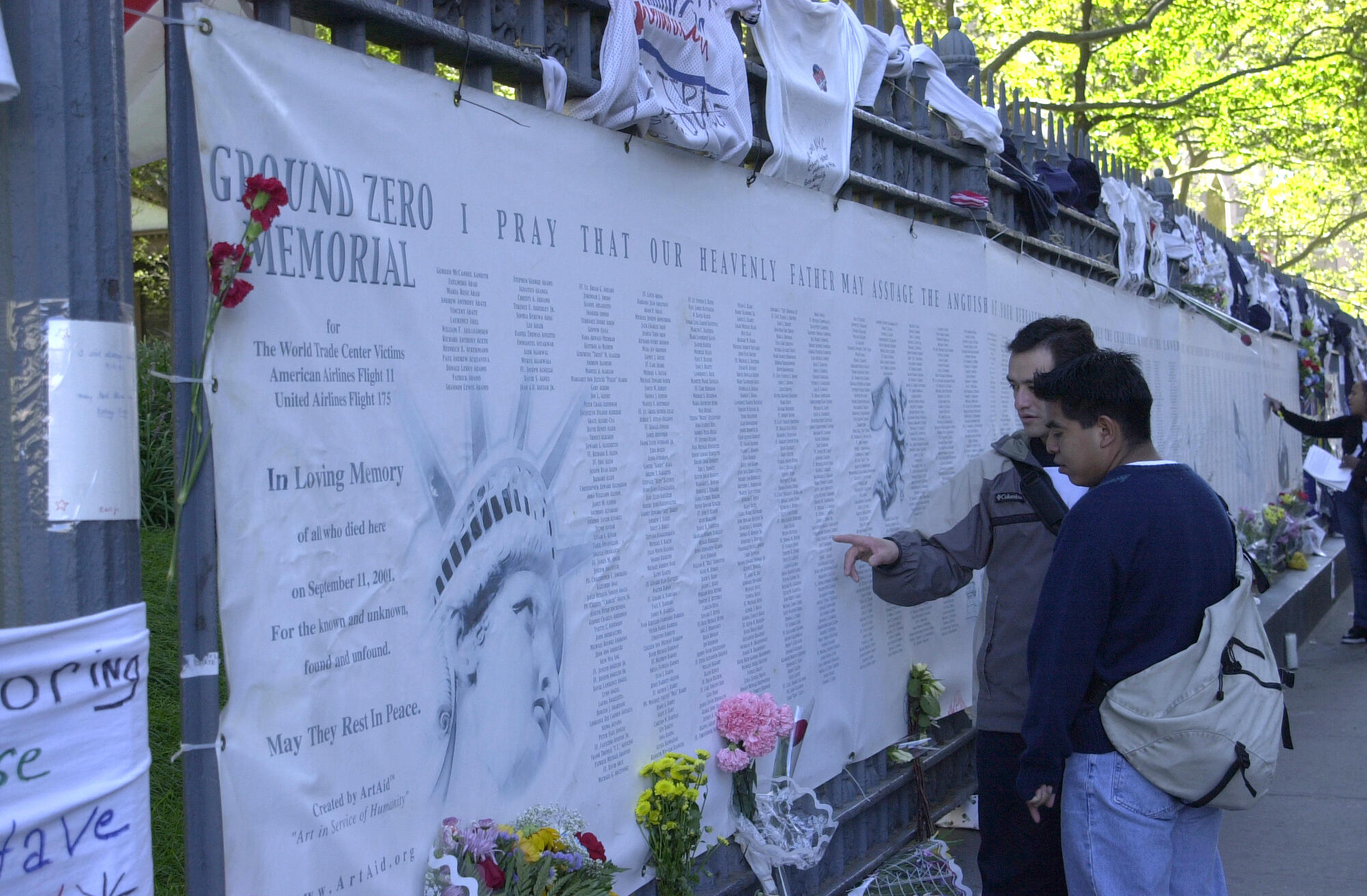 A photo of people reading from a list of thousands of names on a large banner. There are flowers and written messages posted all around.
