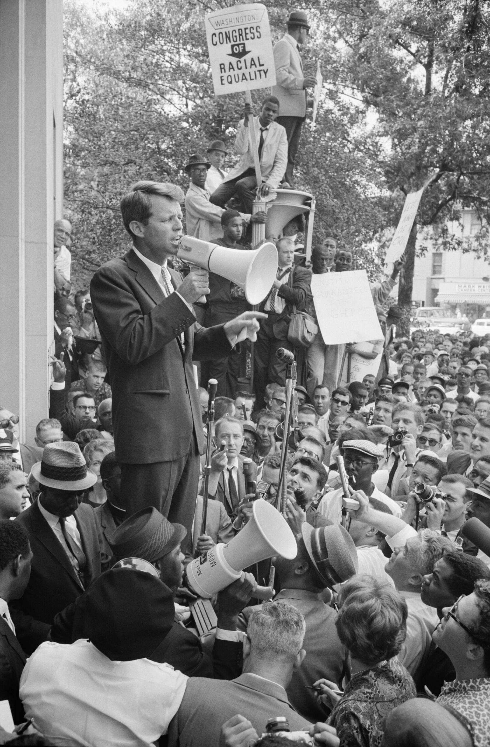 RFK speaking with a megaphone to a crowd. Activists hold up a sign advertising CORE in the background