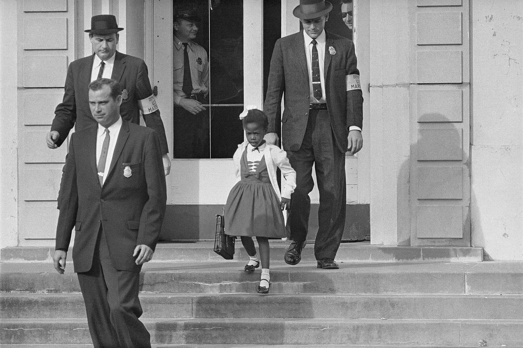 Ruby Bridges wearing her school outfit being escorted down the front steps of her elementary school by US marshals.
