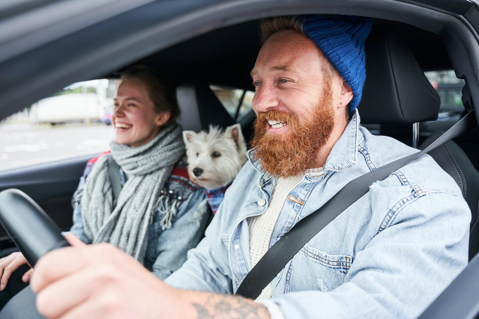A guy with a red beard driving, a women smiling in the passenger seat and a dog in the middle.