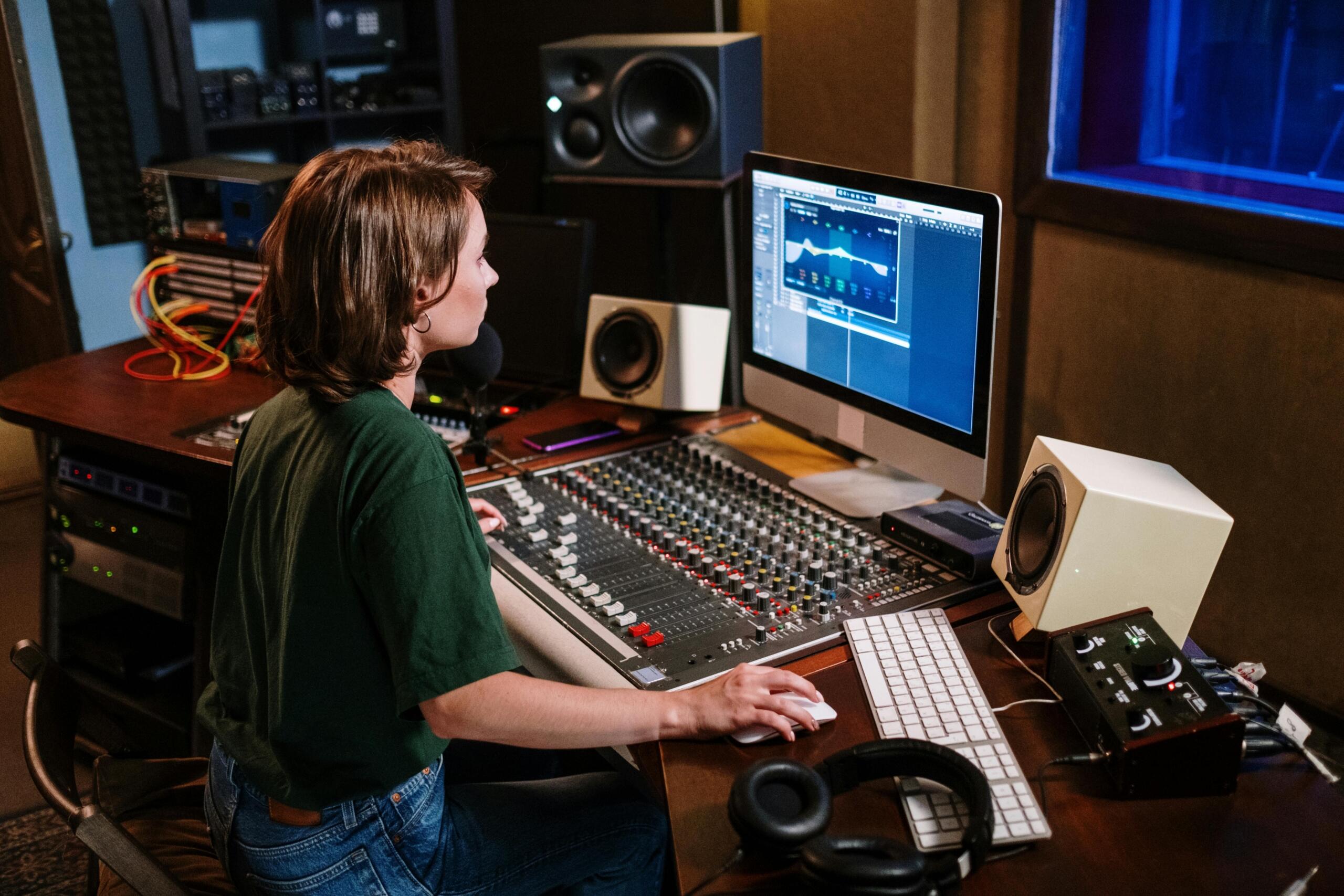 A woman sitting at a desk, adjusting controls on a sound mixer in a recording studio.