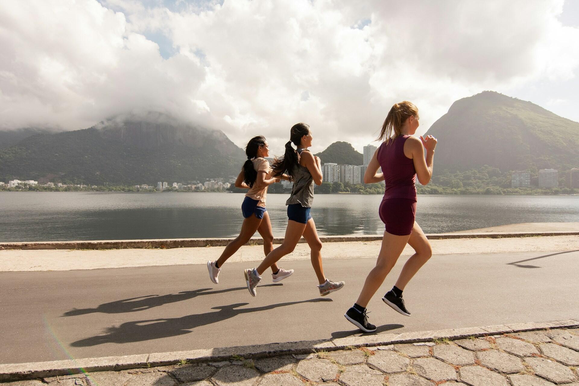 Three girls running next to the water with mountains in the background.