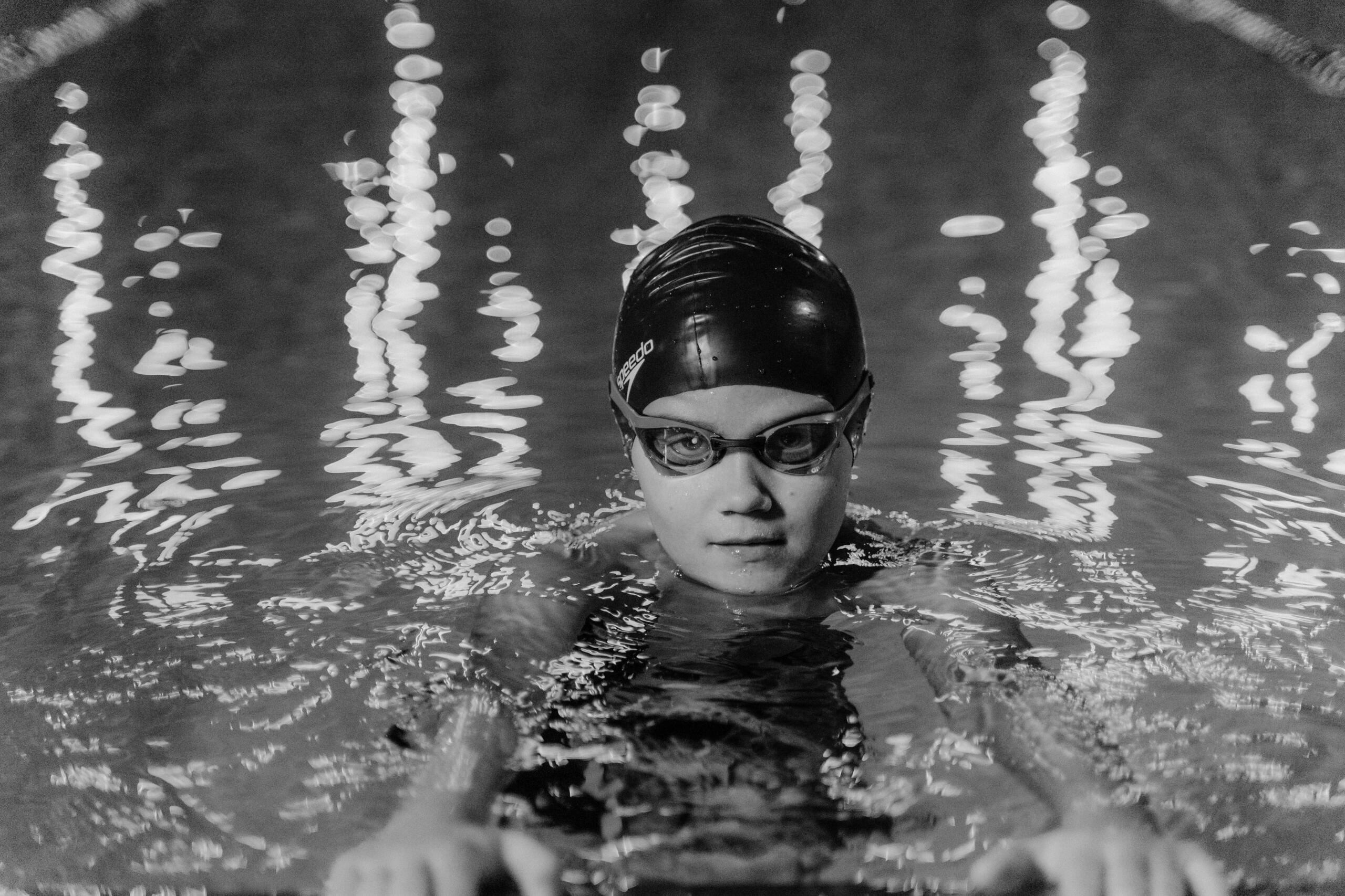 Grayscale photo of a young boy resting his arms on the edge of a swimming pool, looking ahead.