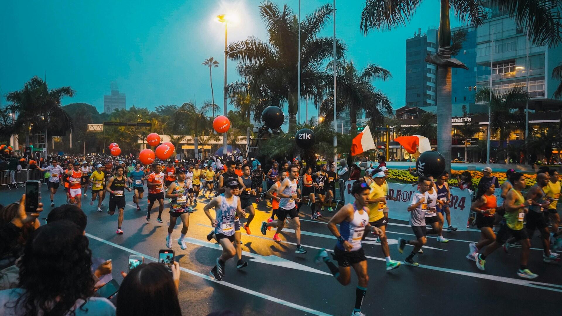 A lively street scene of a marathon with runners wearing bright athletic gear, balloons, and flags under a cloudy sky.
