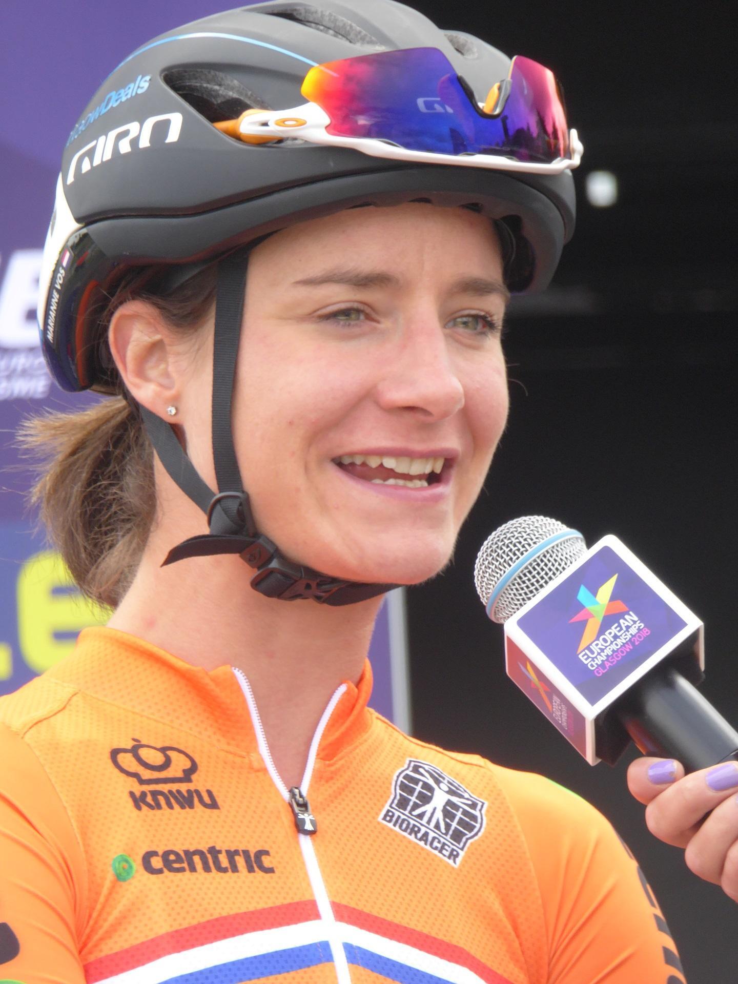 A woman wearing a bicycle helmet and an orange jersey speaks into a microphone. 
