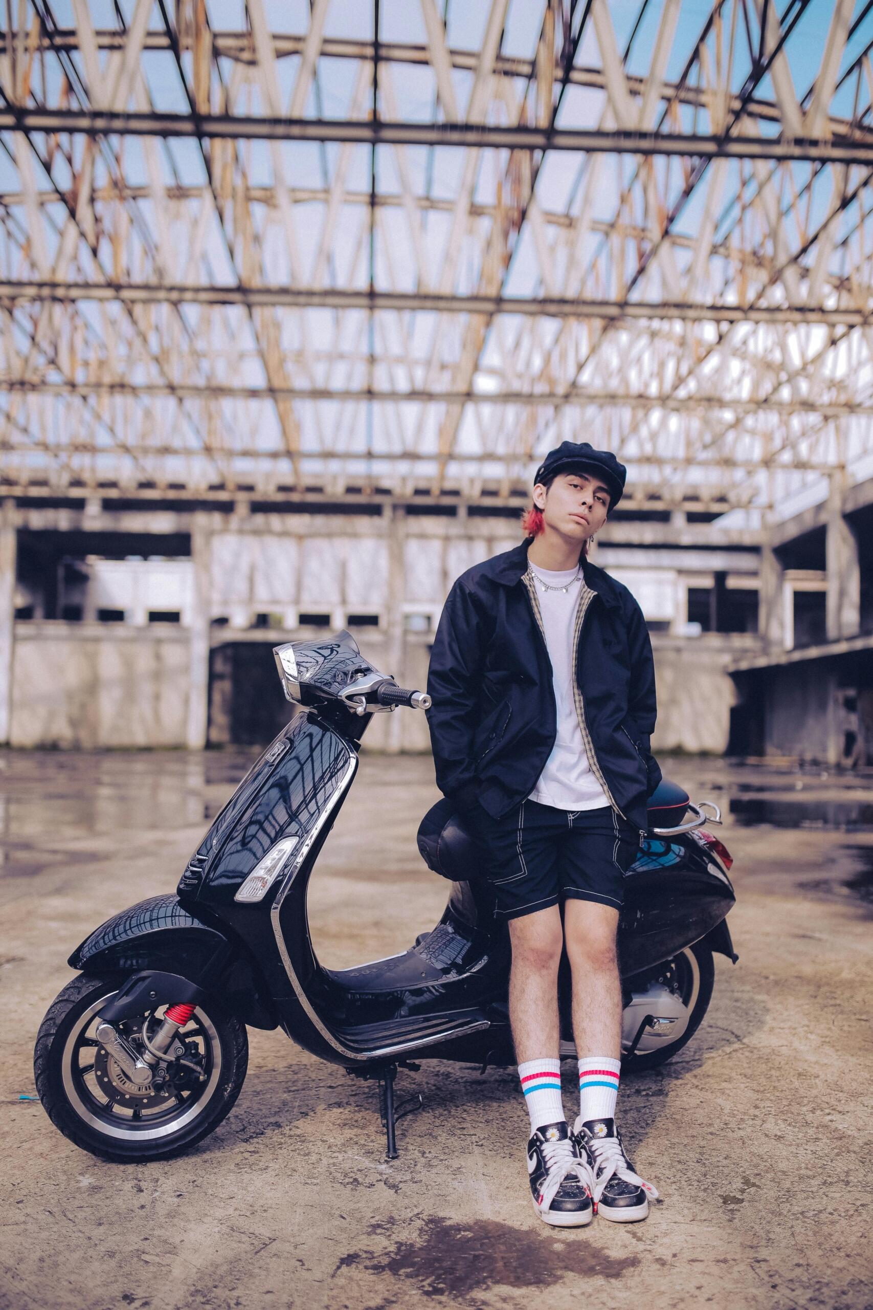 Teenager in a gavroche hat standing next to a scooter in an industrial hall.