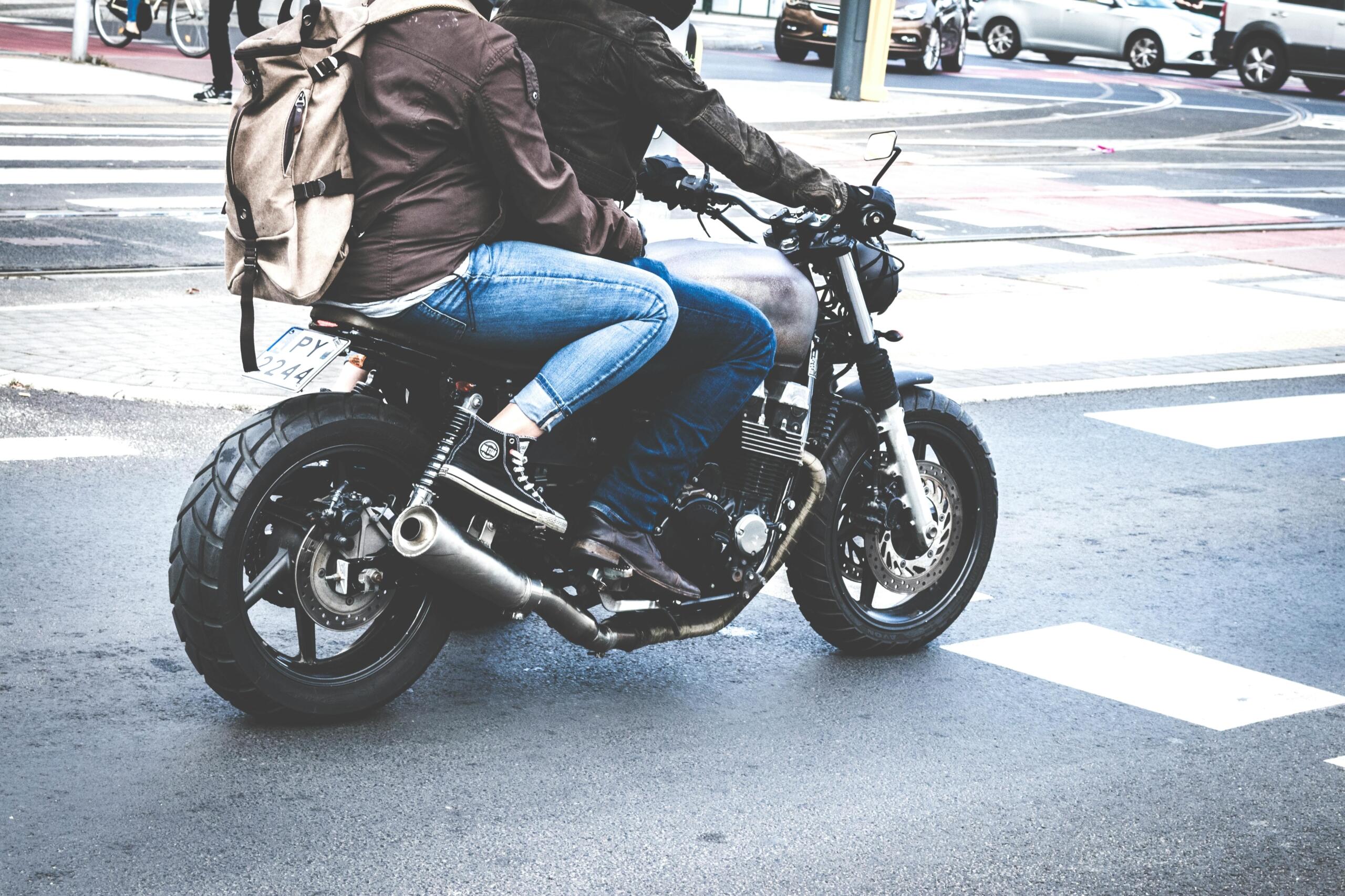 Two people riding a gray café racer motorcycle on the road.