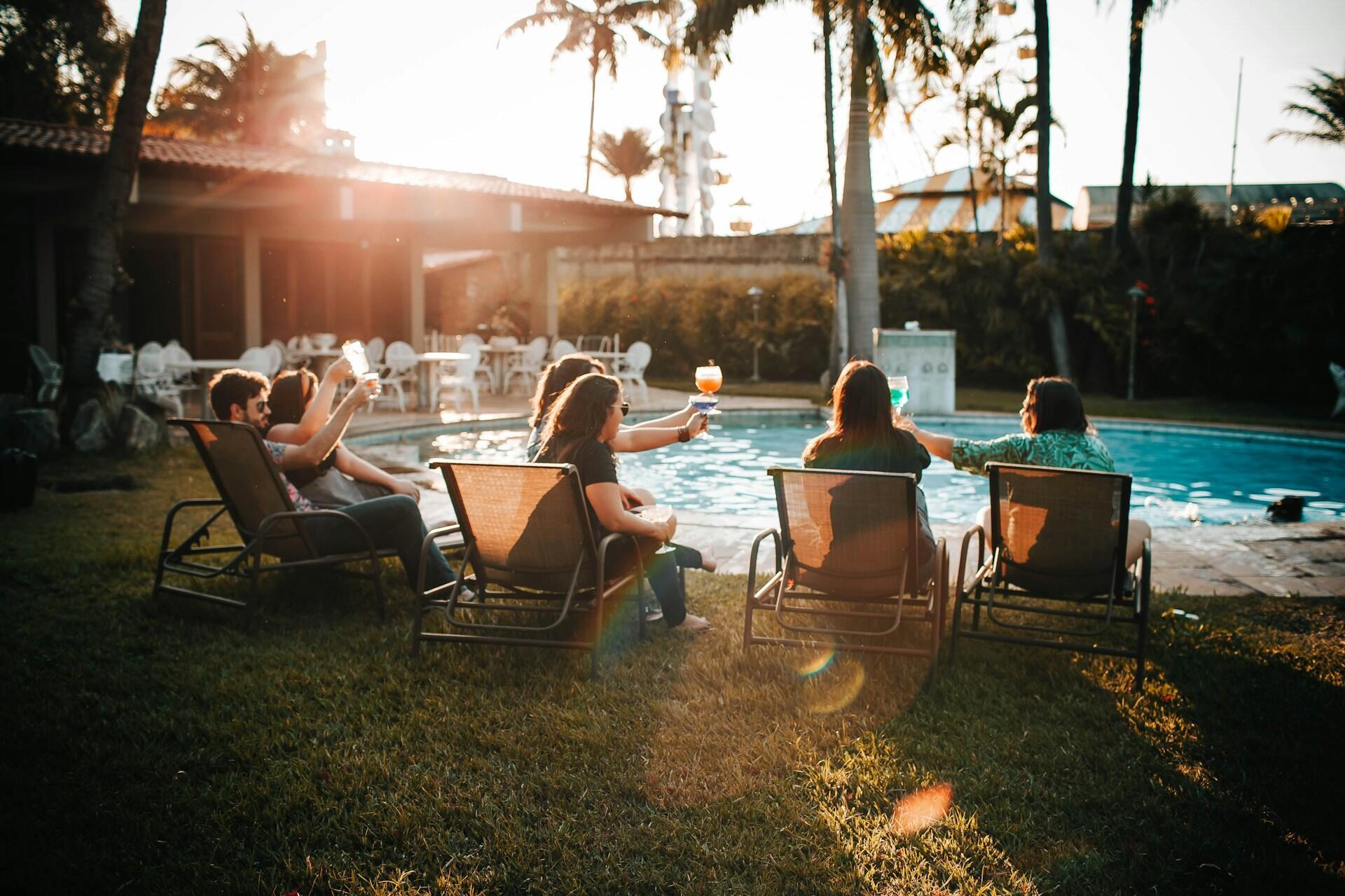 Several people with drinks in their hands sitting on chairs by a pool
