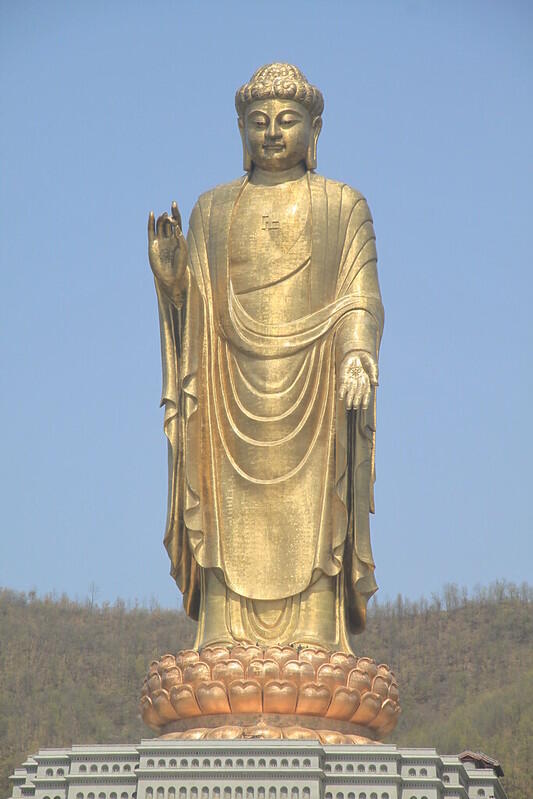 A tall, golden statue of Buddha stands against a clear blue sky, adorned with flowing robes and a peaceful hand gesture. 