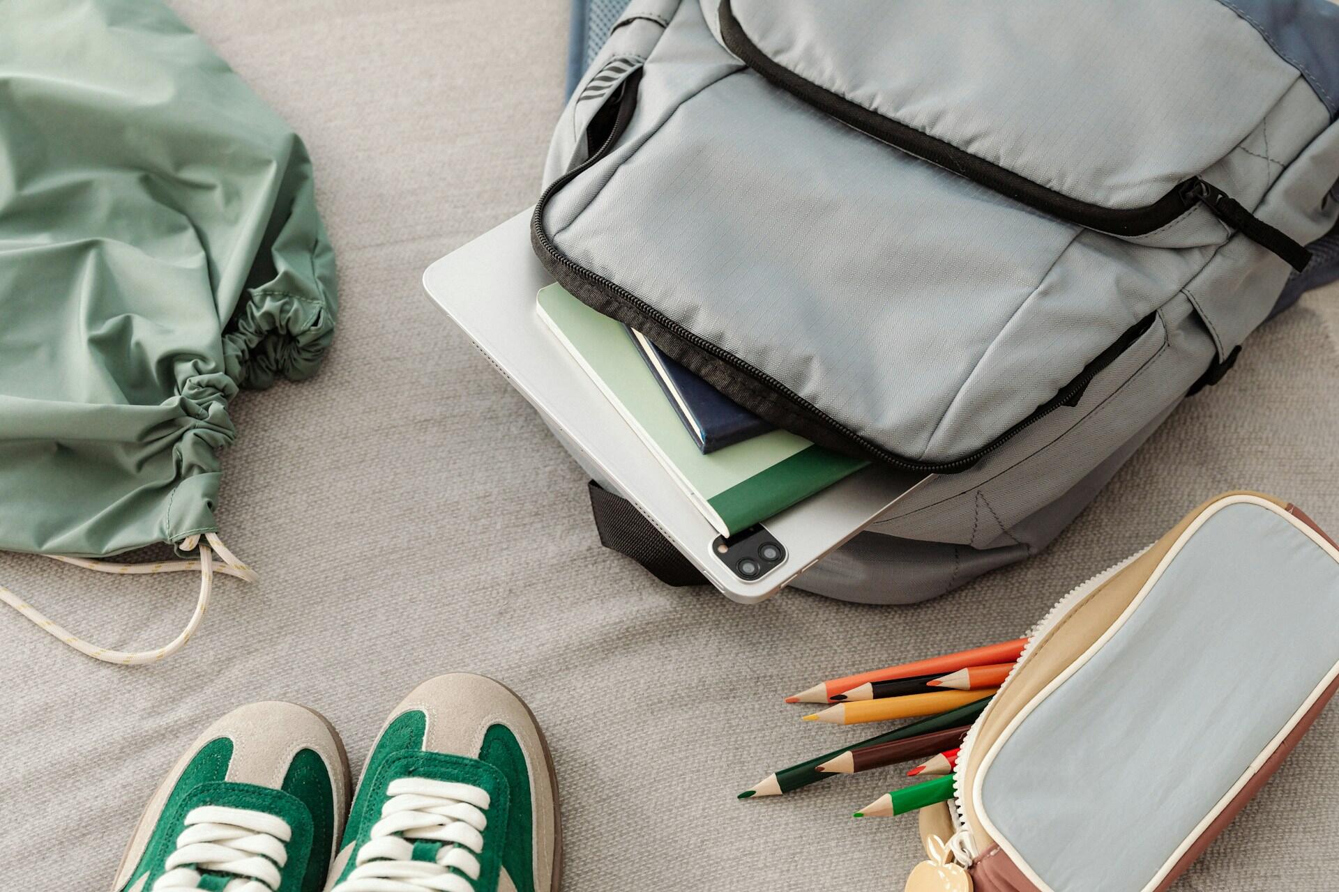a backpack packed with a laptop or tablet and notebooks, near a pencil bag and sneakers.