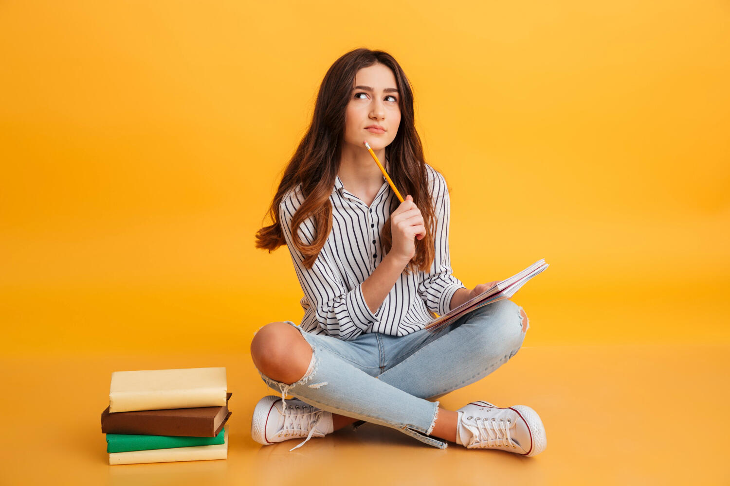 a student sitting on the ground near a pile of books thinking with a pencil and notebook in her hands